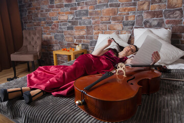 A woman is lying down on a bed in a red dress and looking at music sheets, a cello nearby with a...