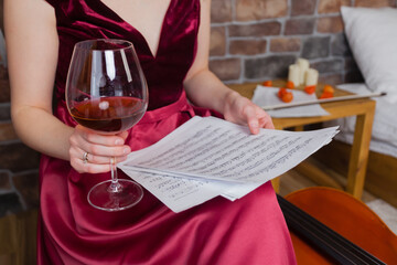 A woman is sitting, looking at musical sheets while holding a glass of wine in her hand.