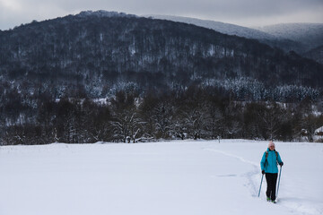 Hiker girl walks along snowy field with mighty mountains in background; hiking in cold winter weather