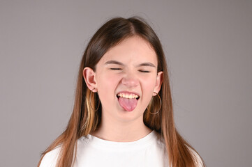 Disgruntled beautiful caucasian girl showing her tongue over gray wall. Teengirl with stylish hoop earrings