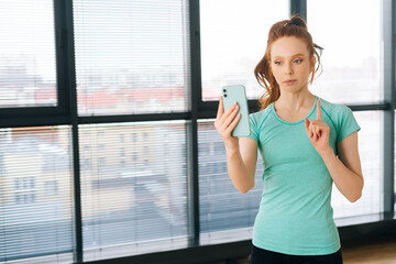 Medium shot of attractive redhead female vlogger holding smartphone, talking to camera shooting vlog, making video call at home talking by virtual mobile video call chat app on background of window.