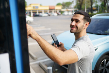 Cheerful happy Asian man using an EV charging application on smartphone to prepare vehicle charging and payment. Modern lifestyle of transportation with sustainability and sustainable energy.