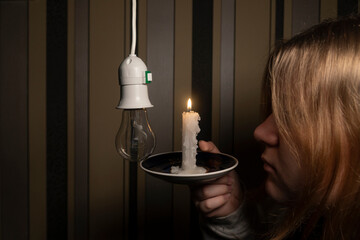 A girl in a dark room holds a burning candle in her hand and looks at the holder with a light bulb (close-up). Blackout. Power outage concept