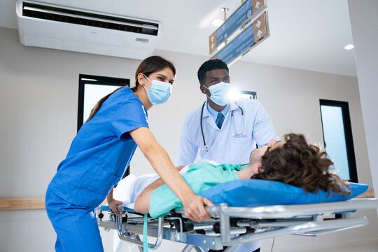 Oblique and low angle view of young African male medicine doctor and young Caucasian female nurse in face masks, urgently pushing Caucasian male patient on emergency stretcher bed at hospital corridor