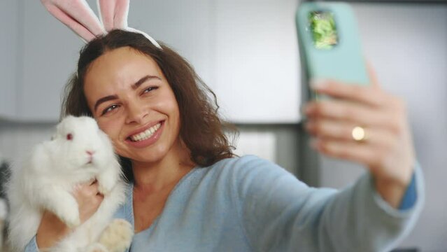 Portrait of the Happy Woman Wearing Bunny Ears Taking a Picture and Playing with Baby Bunny. Smiling Lady Using Smartphone Selfie with Fun Standing in the Kitchen at Home. Happy Easter