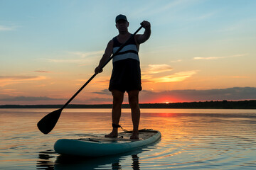 A man in shorts stands on a sapboard with a paddle in the evening at sunset in a lake in the sun glare.