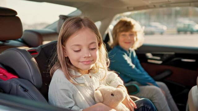 Portrait of cute little blonde girl sitting in back seat of car with beautiful brother looking at camera smiling holding teddy bear. Two lovely kids fastened with safety belts during car trip.