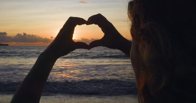 Back view curly hair of woman and hands folded in shape of heart at sunset against background of seascape. Wanderlust, love for travel, summer vacation at sea, holidays. Silhouette, heart-shaped hands