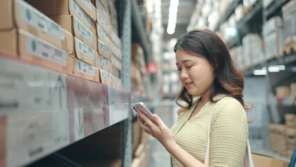 Asian woman shopping on shelves at large warehouse holding and using smartphone scans the QR code for information on furniture at store