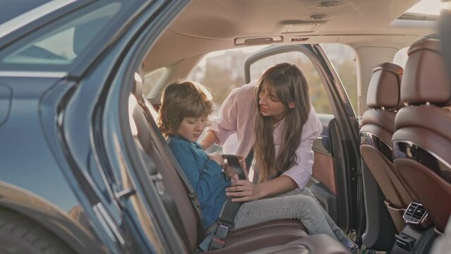 Pretty young caring responsible mother fastening blonde cute little son with safety belts. Happy cheerful boy sitting in back seat of car before family trip bumping fists with father smiling.
