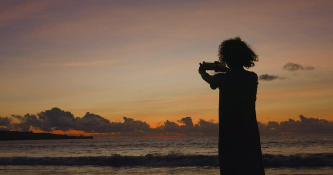 Woman travel blogger, travel photographer pictures dawn on ocean, while waves wash the empty deserted beach. Travel photographer takes photos of early dawn on a smartphone. Silhouette of a curly woman