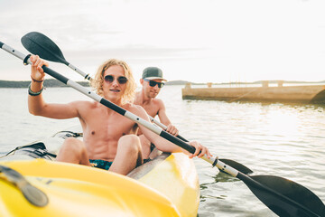 Two smiling rowers father and son on inflatable yellow kayak rowing by the evening sunset rays...