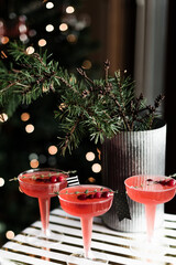 A festive indoor holiday celebration is set next to a lit cozy Christmas tree. Bright red cranberry cocktails complete with decorative flags and topped with a thyme garnish, are illuminated by lights