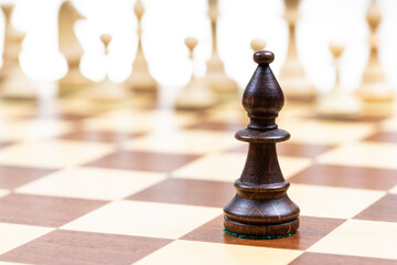 black bishop against white chess pieces in background on wooden chessboard close up (focus on queen) - obrazy, fototapety, plakaty