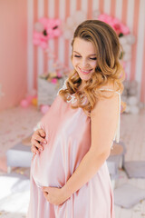 Obraz na płótnie Canvas A beautiful pregnant woman in a light pink dress is smiling and posing on a pink background of a children's room, waiting for a baby.