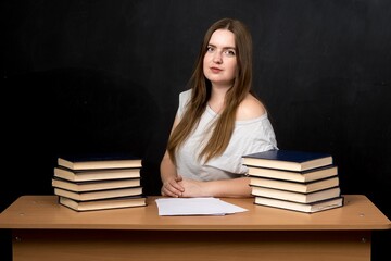 a girl of European appearance, a student or a teacher sits at a desk against a slate wall background near a stack of bookspreparing, serious, academic, project, intelligent, isolated, literature, teen