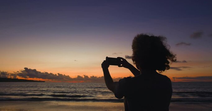 Silhouette Caucasian woman tourist with phone in hands records video in memory of fascinating August sunset and waves washing empty shore. Silhouette of woman recording video from vacation on phone.