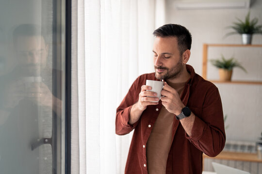 Satisfied Handsome Modern Man Enjoying The Smell Of Morning Coffee, Daydreaming With Eyes Closed While Standing Near Window With White Curtains At Home. Joyful Man With Cup Of Coffee At Home.