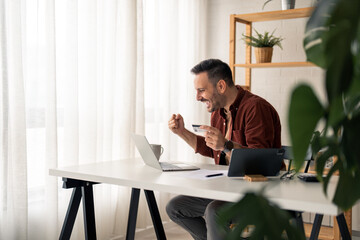 Cheerful businessman in home office feeling excited looking at laptop screen holding credit card, making fist in yes gesture satisfied with online cashback purchase or good electronic banking services