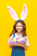 Happy Easter. A little girl holds a basket of colored colored eggs for a spring holiday. A charming child with rabbit ears in a blue sundress on a yellow isolated background