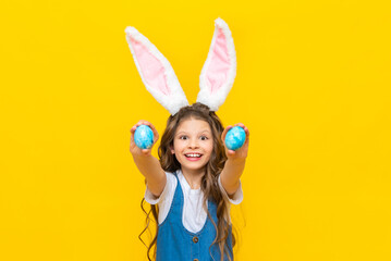 Fototapeta premium Happy spring Easter. A little girl with long hair holds two eggs and stretches them forward, smiling broadly. A charming child with rabbit ears in a blue sundress on a yellow isolated background.