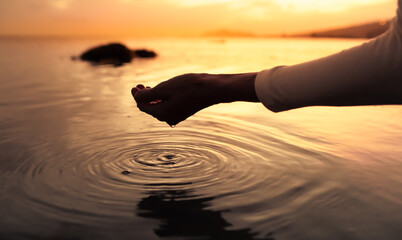 Person's hand holding water with drops making ripples on the surface in a beautiful calm lake setting. Peace in nature concept. 