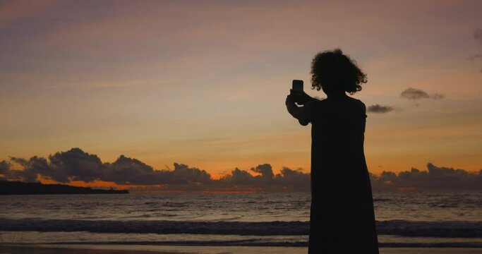 Woman blogger in dress stands by sea against background of orange sunset sky with clouds at very horizon and takes selfie. Woman travel blogger makes beautiful photos of herself, selfies on beach.