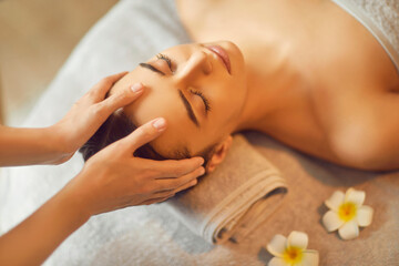 Beautiful young woman receiving facial massage in spa salon. Beautiful girl lying on massage bed on her back with closed eyes enjoying massage. Facial beauty treatment, body care, pampering