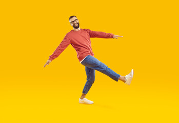 Cheerful and joking young man in youth casual clothes takes step on orange background in studio. Stylish bearded guy in sweatshirt, jeans and sneakers takes step while looking at camera. Full length.