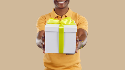 Close up of festive gift box in hands of joyful dark-skinned man on beige background. White box with yellow ribbon in hands of unrecognizable smiling man. Congratulations concept. Web banner.