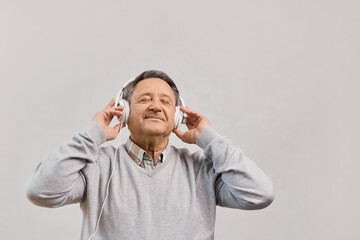 Senior man with closed eyes against a gray background wearing headphones listens music, having nostalgic mood, enjoys songs of his youth, older generation using modern technology concept, copy space
