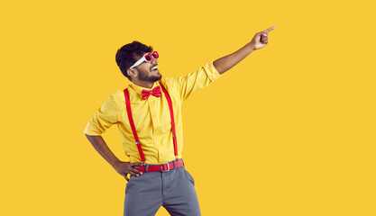 Young funny Indian man with smile points to copy space for your promotional offer and invites you to take part in promo action dressed in bright shirt with bow tie stands in yellow studio - obrazy, fototapety, plakaty