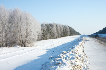 Fototapeta na wymiar A winter highway going over the horizon. Along the road is a beautiful white grove in hoarfrost. A lone car rides along the road, snow cleared to the side of the road. It's a frosty day.