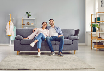Happy smiling young couple are sitting together on cozy sofa at home in the living room, looking into the camera. Sense of harmony in relationships, care, joy, positivity, love, romance, satisfaction.