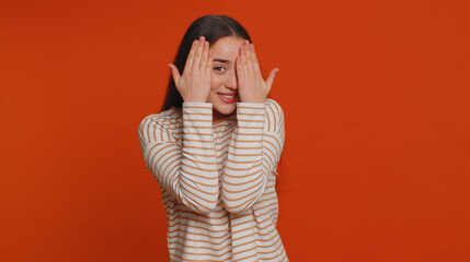 Nosy curious young woman closing eyes with hand and spying through fingers, hiding and peeping, binocular gesture, exploring way, seeking something in distance. Girl isolated on red studio background