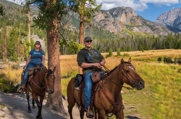Smiling young man and woman riding horses in the forest of Rocky Mountains, Colorado, in fall; meadow and mountain range in background