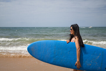 South American woman, young and beautiful, brunette with sunglasses and swimsuit, coming out of the water holding a blue surfboard. Concept sea, sand, sun, beach, vacation, surf, summer.