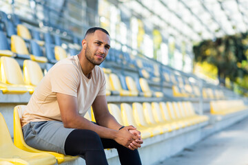 Portrait of a young handsome African-American sportsman sitting on a chair on a tribune in a stadium. He looks at the camera, smiles.