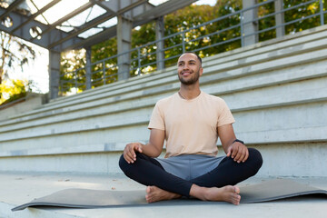 Fototapeta na wymiar Morning meditation. A young African American man is sitting on a mat in a stadium in the lotus position and doing yoga. Looking ahead, smiling, relaxed.