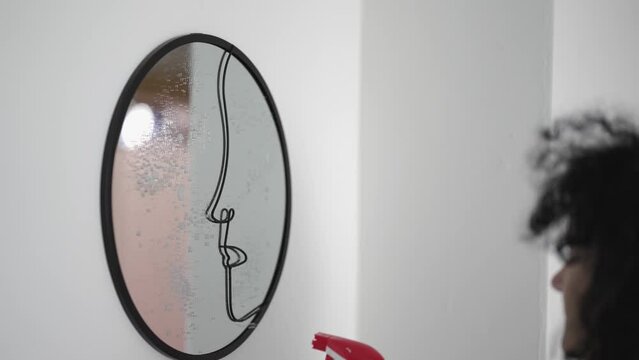Housework and housekeeping concept - Curly hair woman cleaning face shaped mirror with rag at home, slow motion