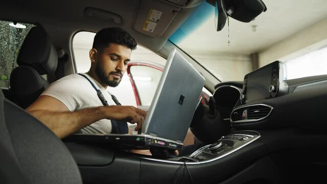 Young bearded man car technician mechanic sitting inside red car repairing problem of engine, during system checking detail, using laptop computer for maintenance and fixing in garage.