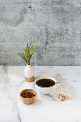 Small bud vase with a palm leaf sits next to a ramekin of roasted coffee beans on a tray. It also features black coffee in a teacup with sugar cubes.