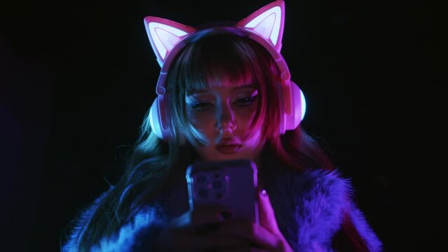 Cute gamer girl wearing headphones with luminous cat ears and playing a game on her phone