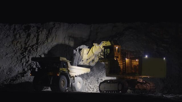 Earth mover loading dumper truck with rocks in open pit quarry mine at night, slow motion