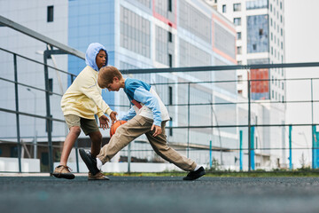 Two adolescent intercultural schoolboys in activewear playing basketball on playground while trying to hold the ball during sports game