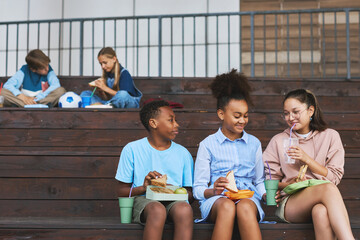 Group of three happy multicultural school friends having sandwiches and soda for lunch while...