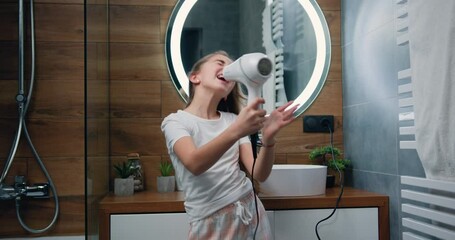 Beautiful blond little girl in bathrobe singing a song and dancing with a hair dryer. Child sings and dances in a shower after taking shower in bathroom.