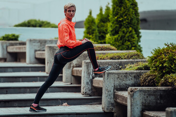 Fit attractive woman in sportswear stretching one leg before jogging on the footpath outdoor in summer among greenery. Workout, sport, activity, fitness, vacation and training concept.