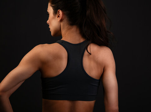 Serious female sporty muscular with ponytail doing stretching workout the shoulders, blades and arms in sport bra, standing on dark grey background with empty copy space. Back view.