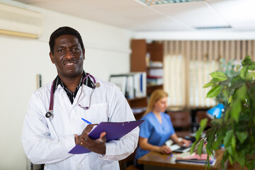 Portrait of smiling african american doctor wearing white coat meeting patient in medical office, filling out medical form at clipboard - obrazy, fototapety, plakaty
